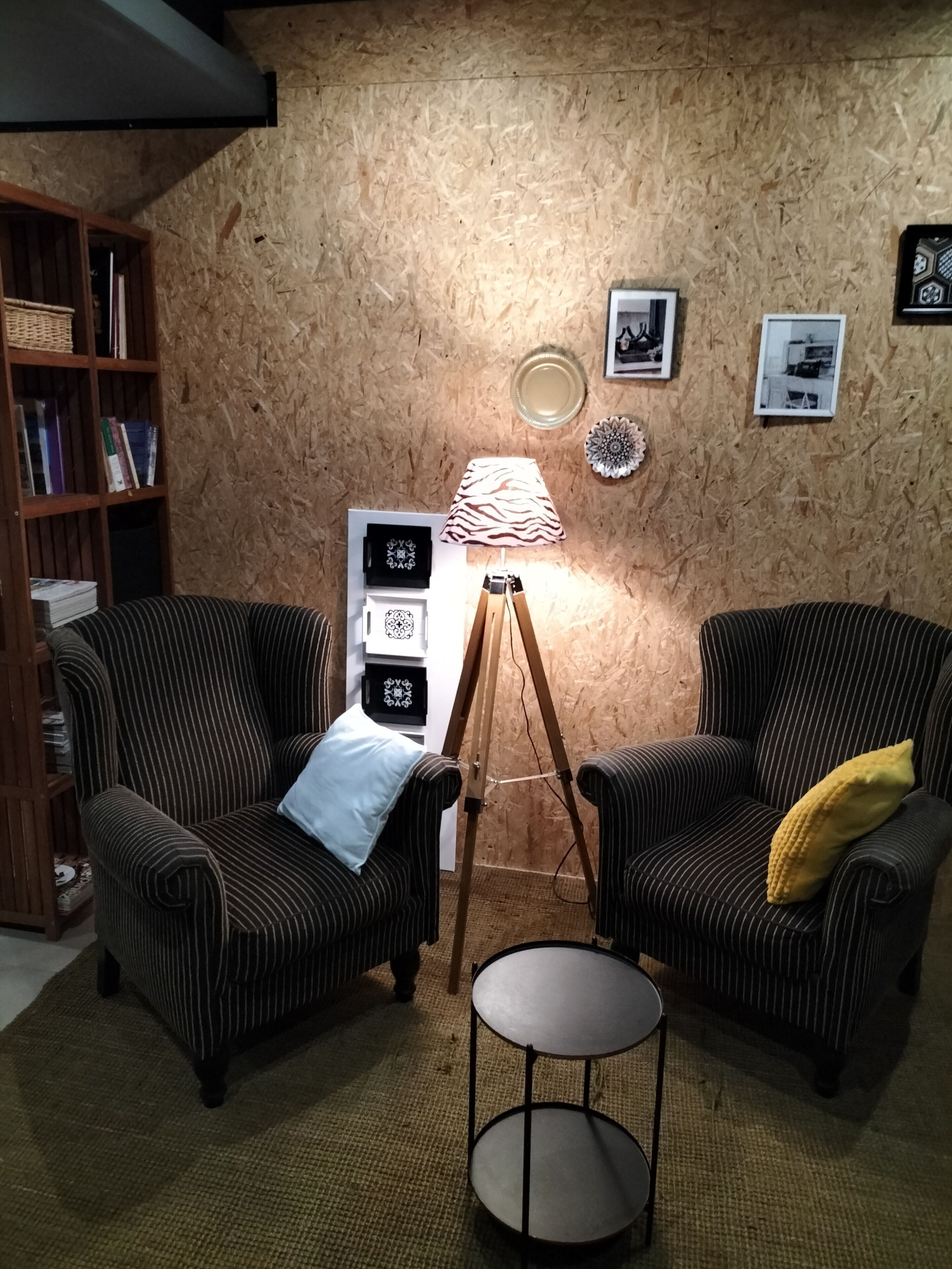 A room with two striped armchairs, a small round table, a tripod lamp, a bookshelf, and framed pictures on a wooden wall.