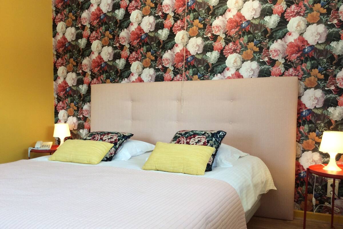 A hotel room with a floral wallpaper, a tufted headboard, and a bed with two pillows.