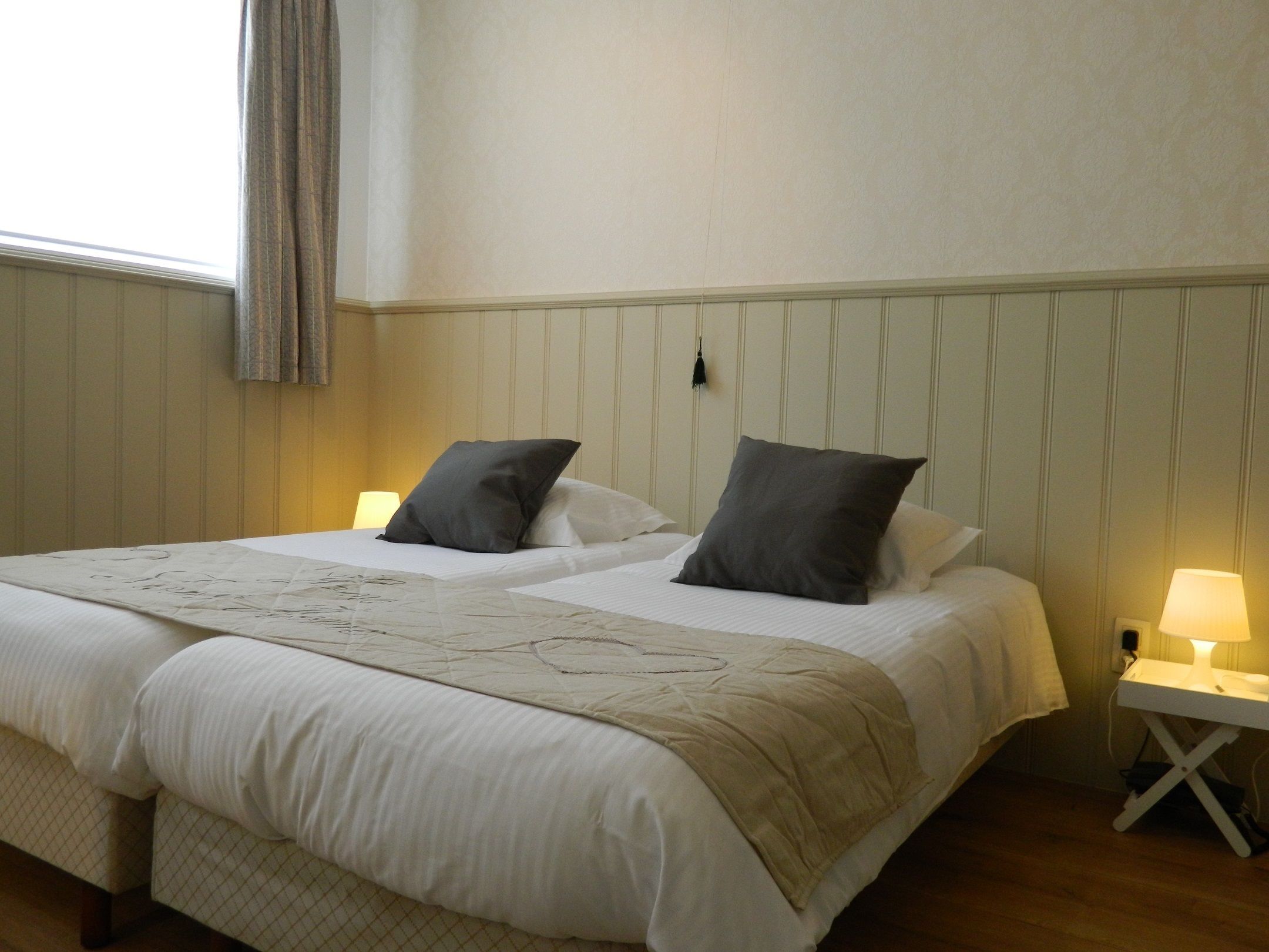 A neatly made bed with a white duvet and two pillows, a bedside table with a lamp, and a window with curtains.