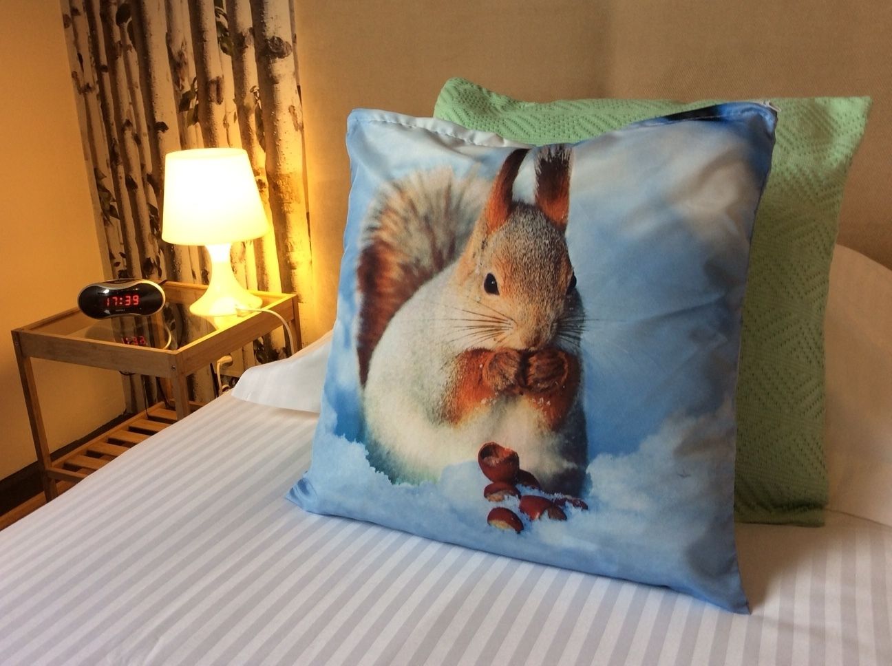Una cama con un almohadón que muestra una ardilla y bellotas, una lámpara de noche y un reloj digital sobre una mesita de noche de madera.