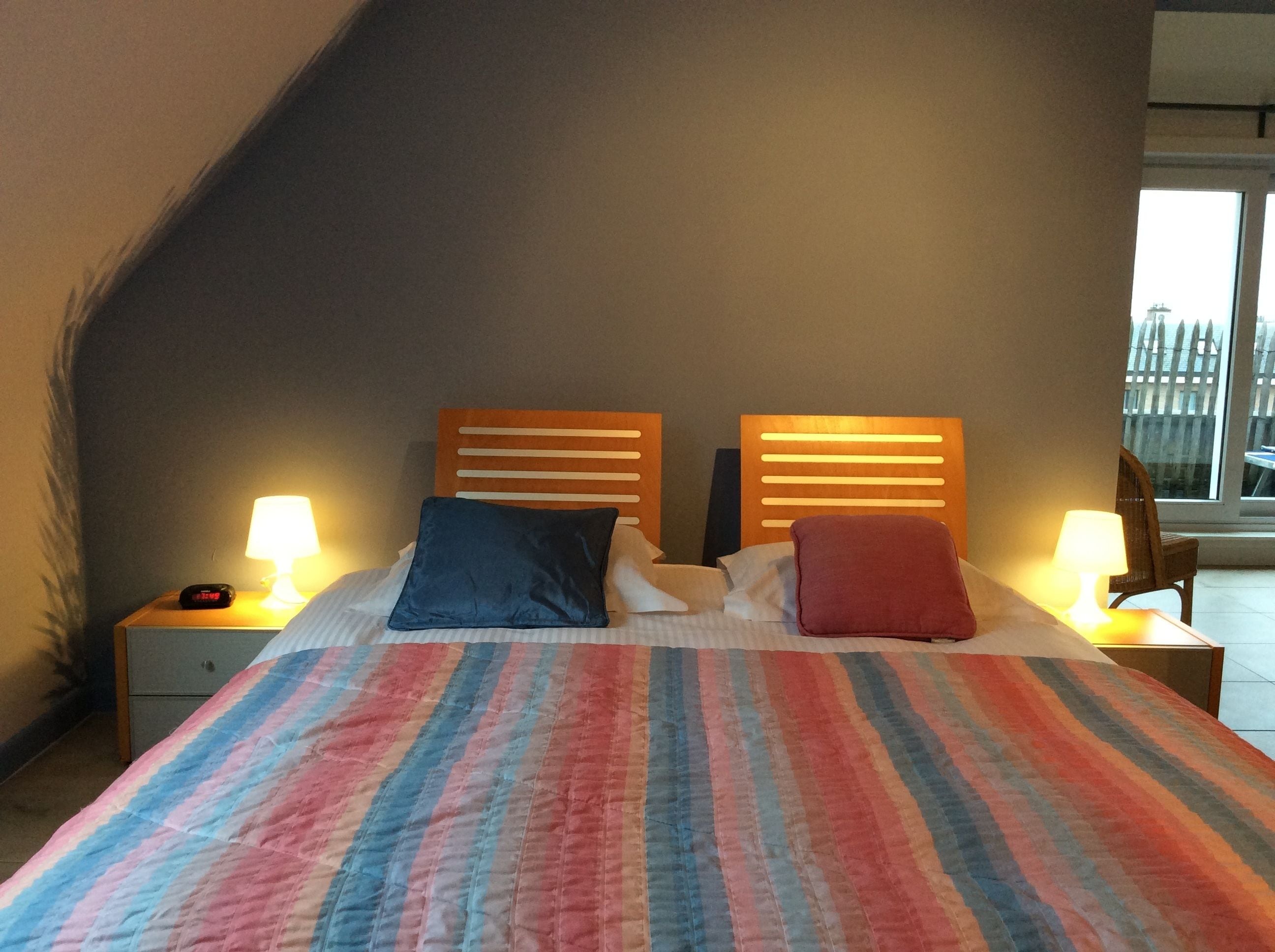 A bedroom with a sloped ceiling, a bed with a striped blanket, two bedside lamps, and a view of a balcony with a fence.