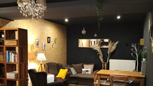 A cozy living room with a sofa, armchair, and a dining table. There is a bookshelf and a chandelier. - Hotel Butler (Zuienkerke)