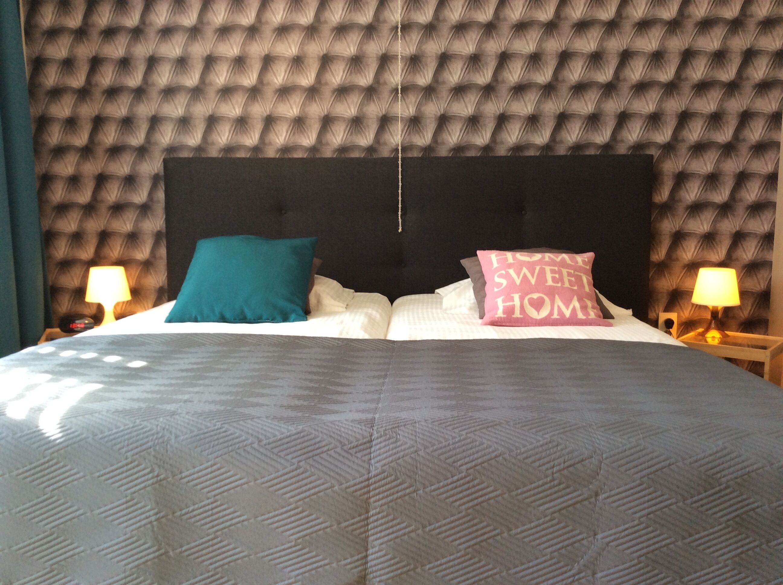 Ein Hotelzimmer mit gebogenem Kopfstück, einem großen Bett mit gemusterten Bettbezügen, zwei Nachttischlampen und einem Kissen mit der Aufschrift 'HOME SWEET HOME'.