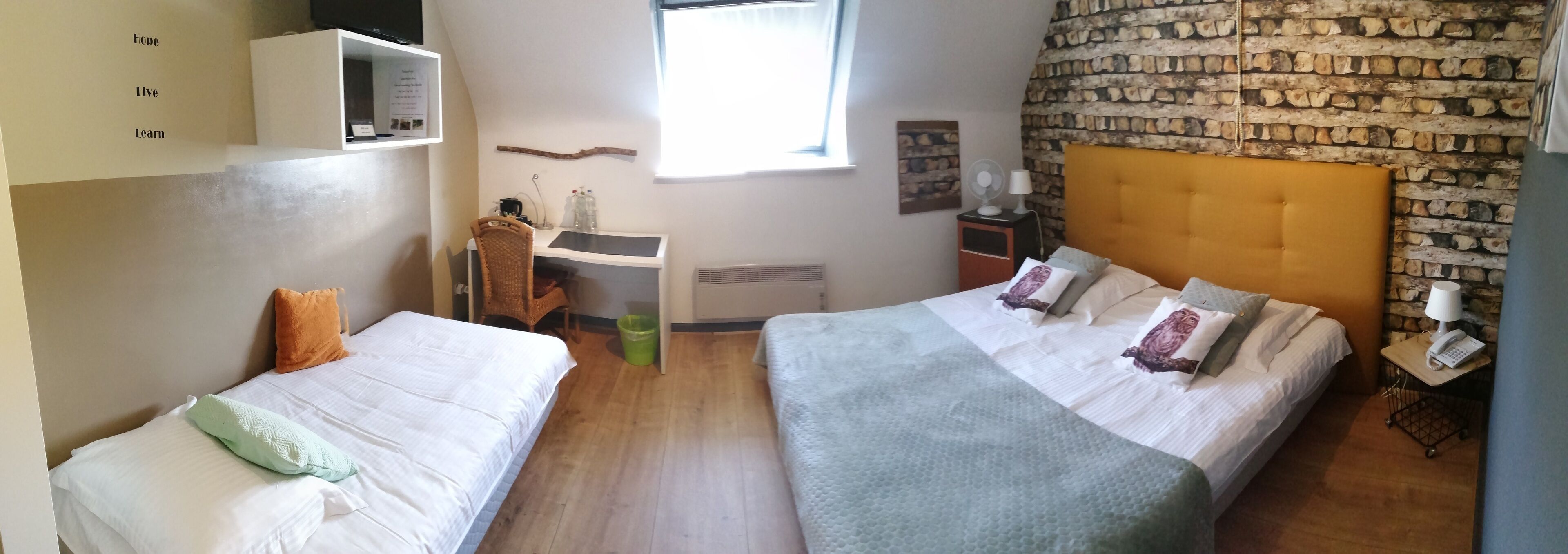 Una habitación con dos camas, un escritorio, una silla, una mesita con lámpara, un ventilador y una pared de piedra.