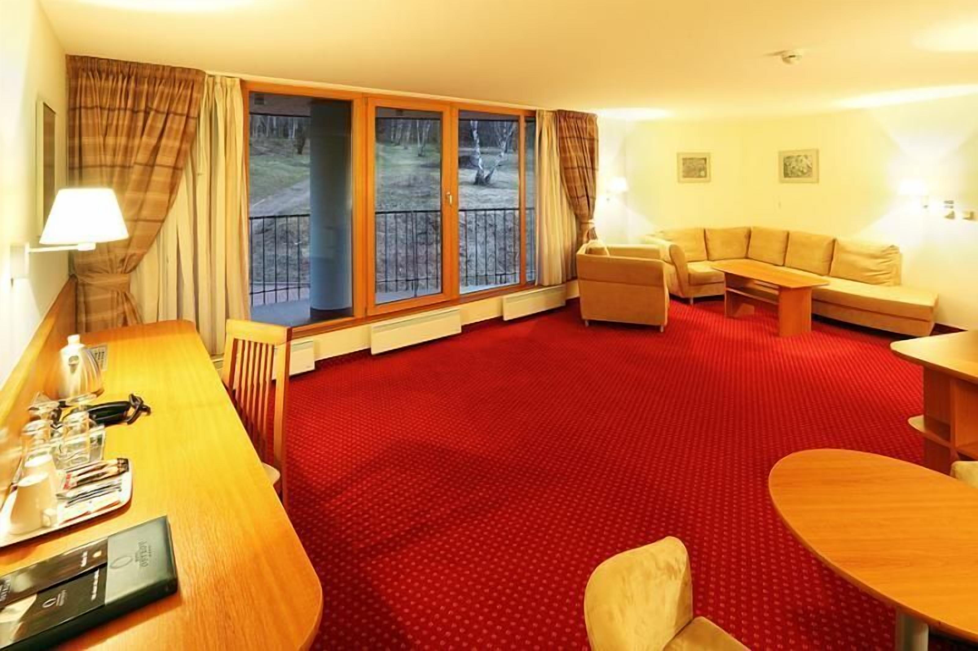 Quarto de hotel com carpete vermelho, escrivaninha de madeira com luminária, sofá, mesa pequena e vista para uma varanda com grade.