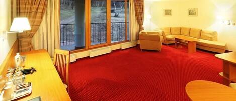 A hotel room with a red carpet, a wooden desk with a lamp, a sofa, a small table, and a view of a balcony with a railing.