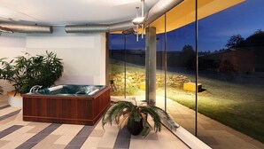 A modern indoor space with a hot tub, potted plants, and a view of a stone wall and grassy area outside. - Hotel Ostrov (Tisa)