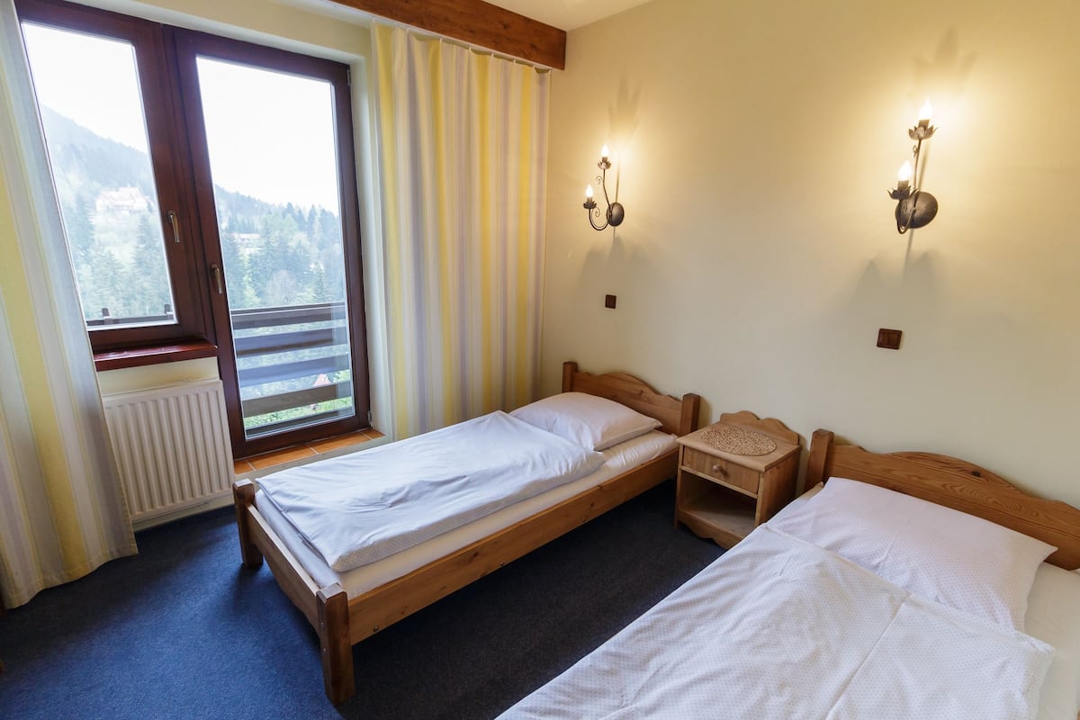Ein Zimmer mit zwei hölzernen Betten, einem fenster mit Vorhängen und einer Wandleuchte.