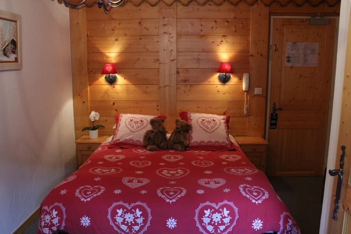 A cozy room with a wooden headboard, two bedside lamps, a bed with a red heart-patterned bedspread, and two teddy bears on the pillows.