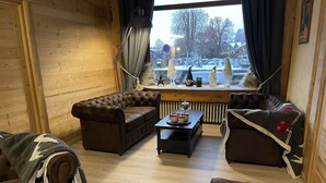A cozy living room with a brown leather sofa, a matching armchair, and a small table with a teapot and cups. A radiator is visible under the window, and there are decorative items on the windowsill. - Hôtel les Glaciers (Samoëns)