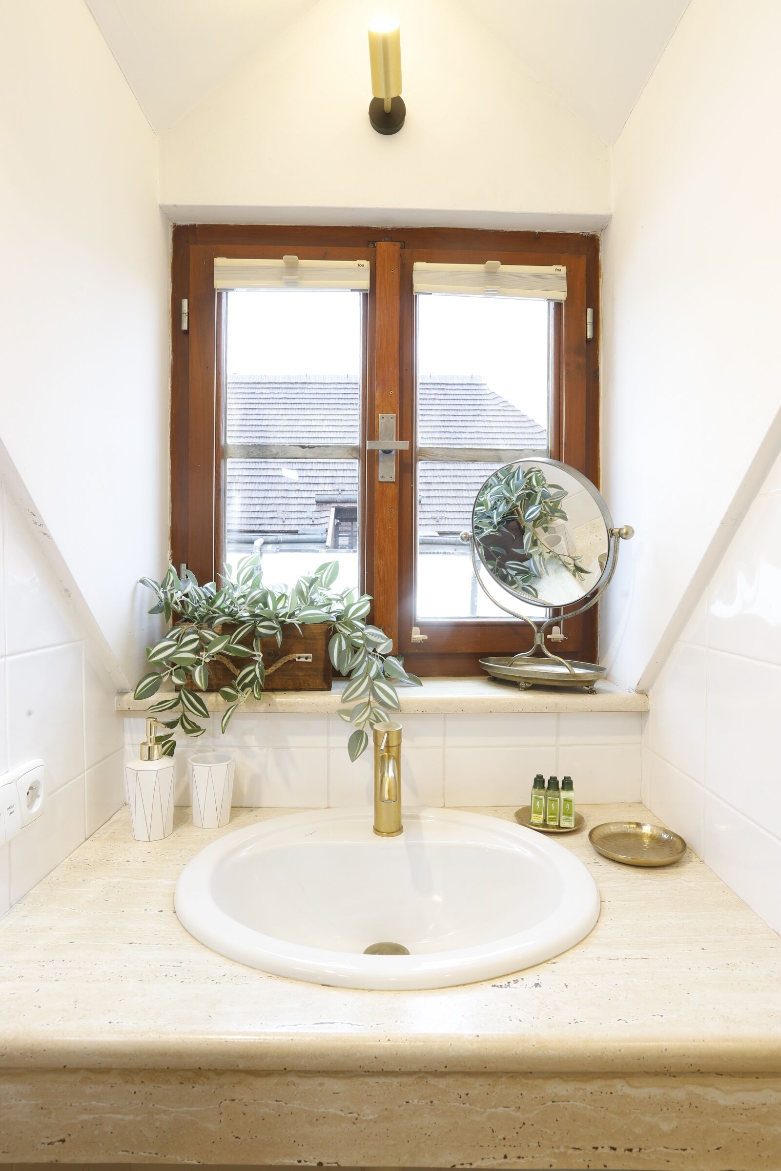 Une salle de bain avec un lavabo blanc, un miroir, une fenêtre avec des stores et une plante en pot.