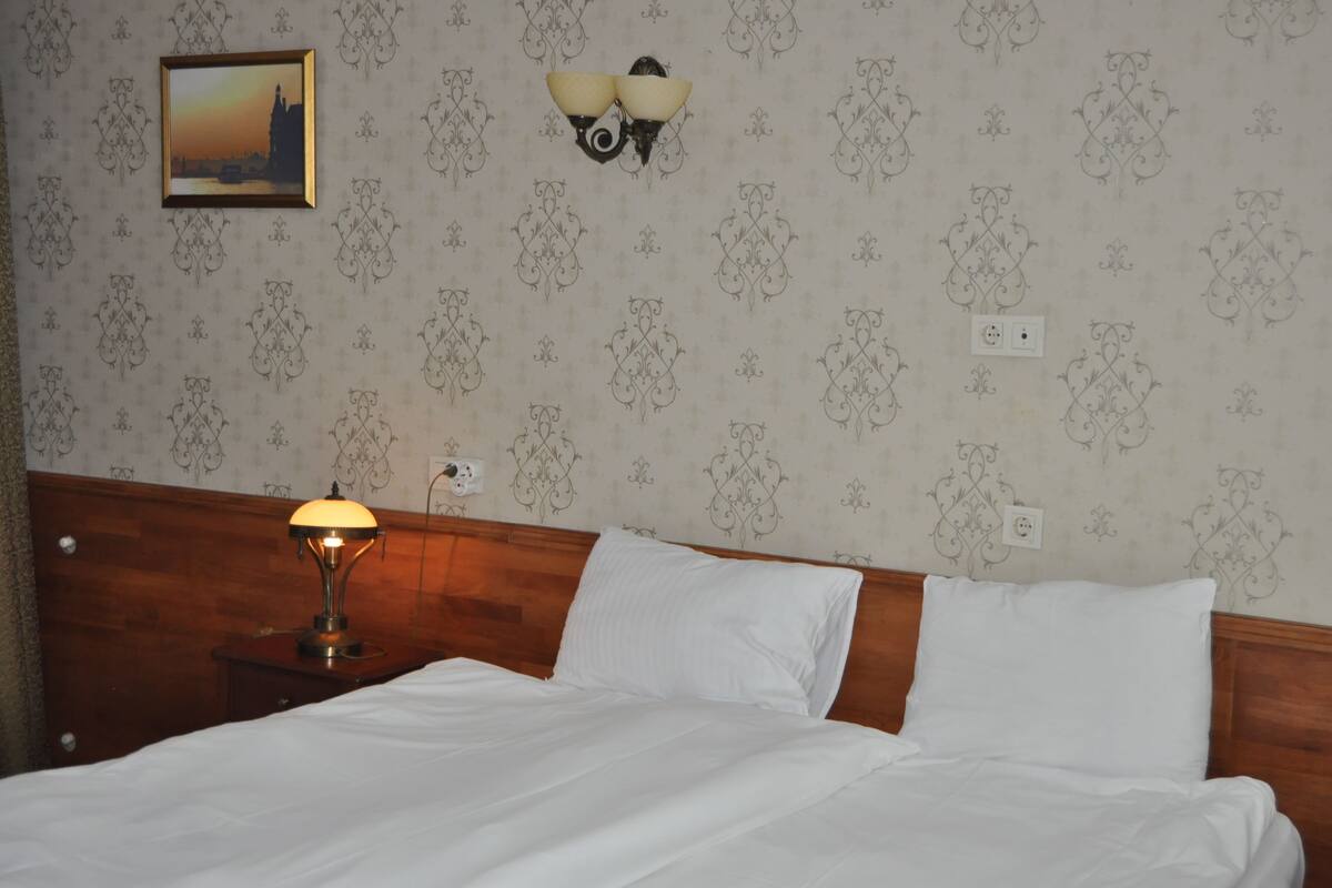 A neatly made bed with white linens, a bedside table with a lamp, a wall-mounted picture frame, and a wall sconce.