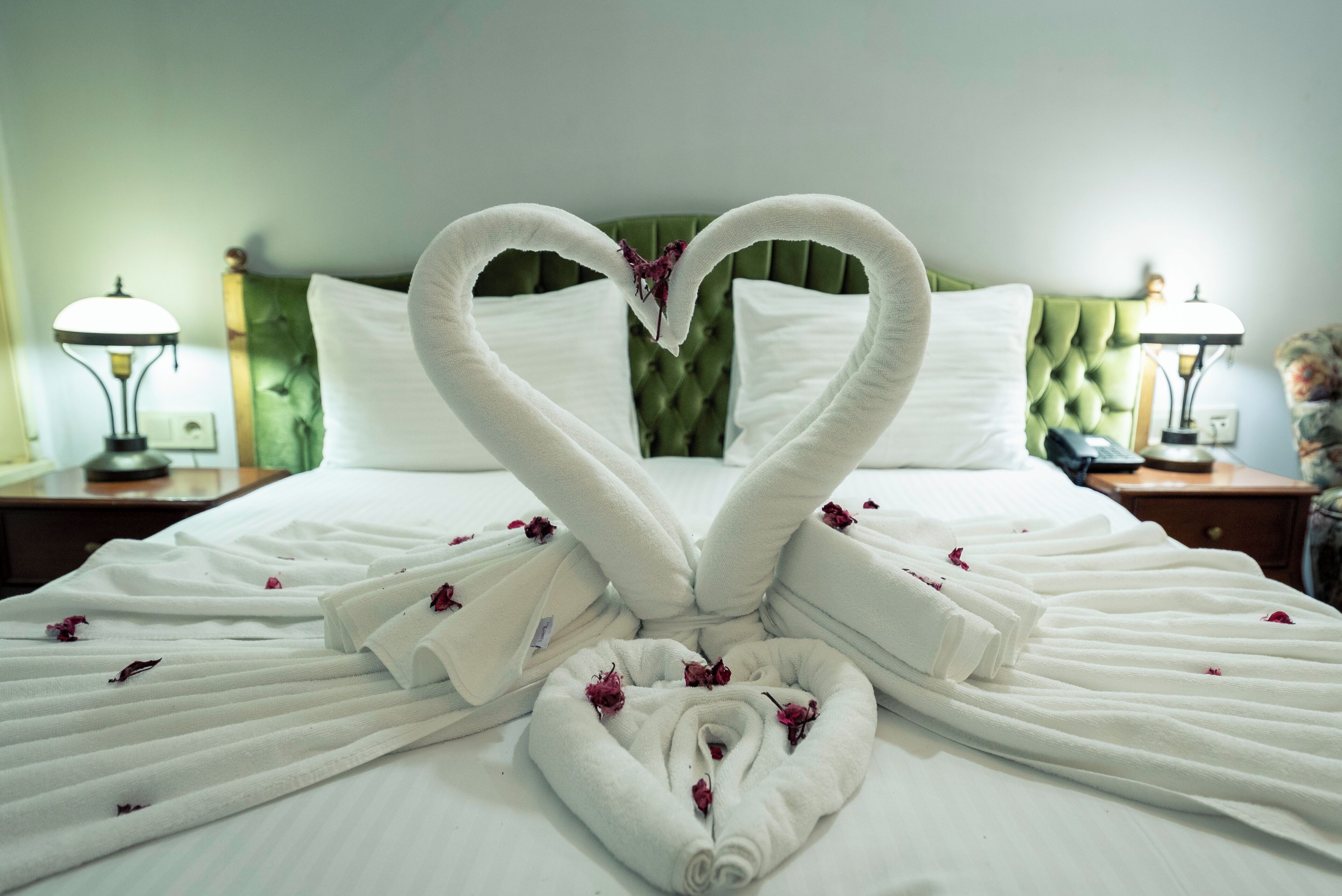 A neatly made bed with towels arranged in a heart shape, a green upholstered headboard, and two bedside lamps.