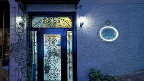 Der Eingang des Hotels Basileus mit einem dekorativen Eisengitter, einer Fußmatte und einem Neonlichtschild an der Wand.