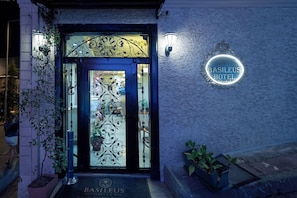 Der Eingang des Hotels Basileus mit einem dekorativen Eisengitter, einer Fußmatte und einem Neonlichtschild an der Wand.