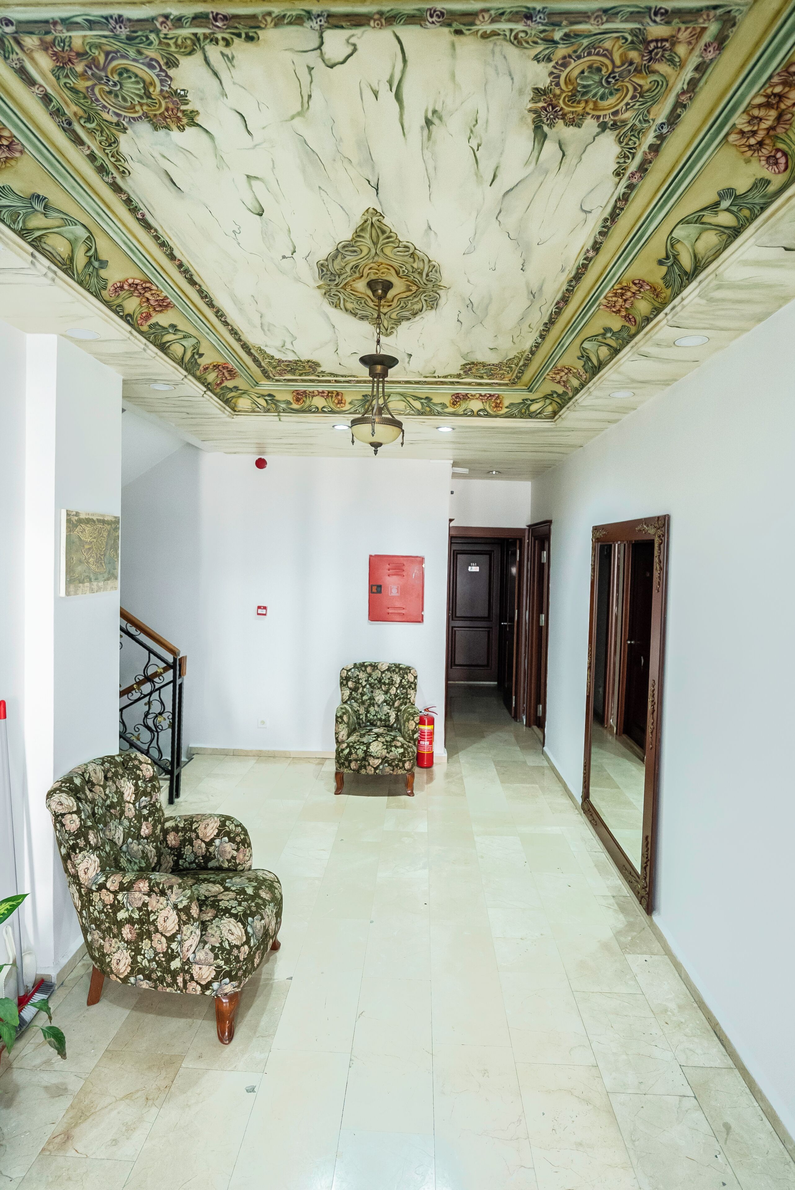A spacious room with a high ceiling, a floral-patterned armchair, a fire extinguisher, and a door leading to another area.