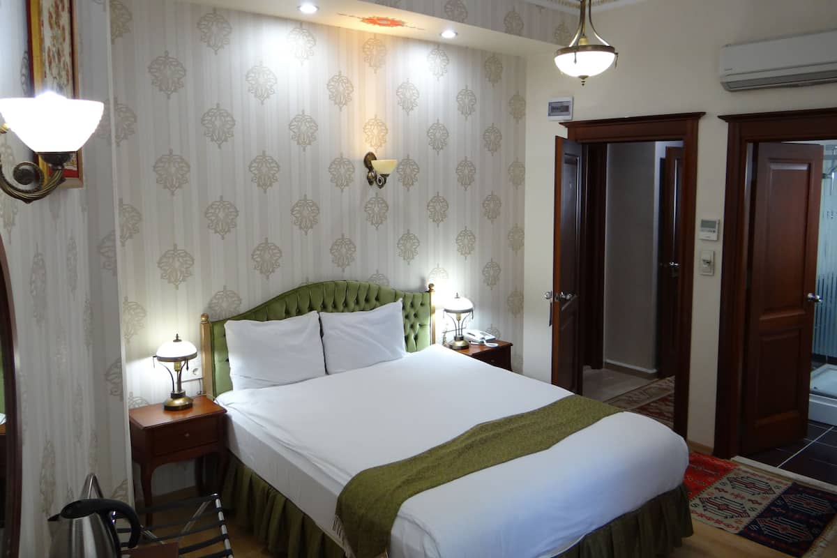 A hotel room with a large bed, two bedside lamps, a green throw blanket, a wooden nightstand, and a wall-mounted lamp.