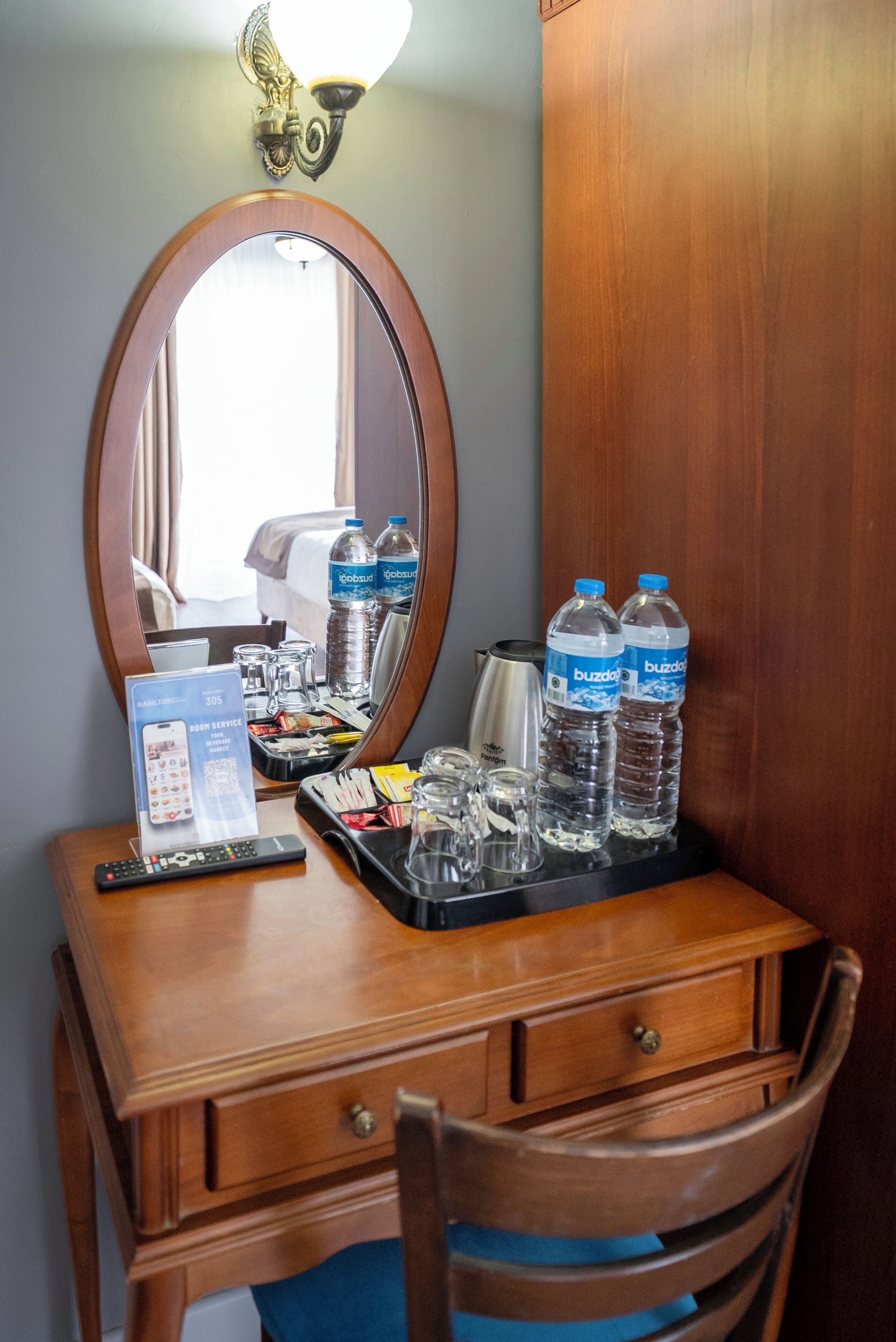 A hotel room with a wooden desk, a mirror, a tray with water bottles, a remote control, and a minibar.