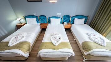 A hotel room with three single beds, each with a white duvet and a towel with a heart design. There are wooden nightstands with lamps and a telephone. The room has a blue headboard and a green curtain.