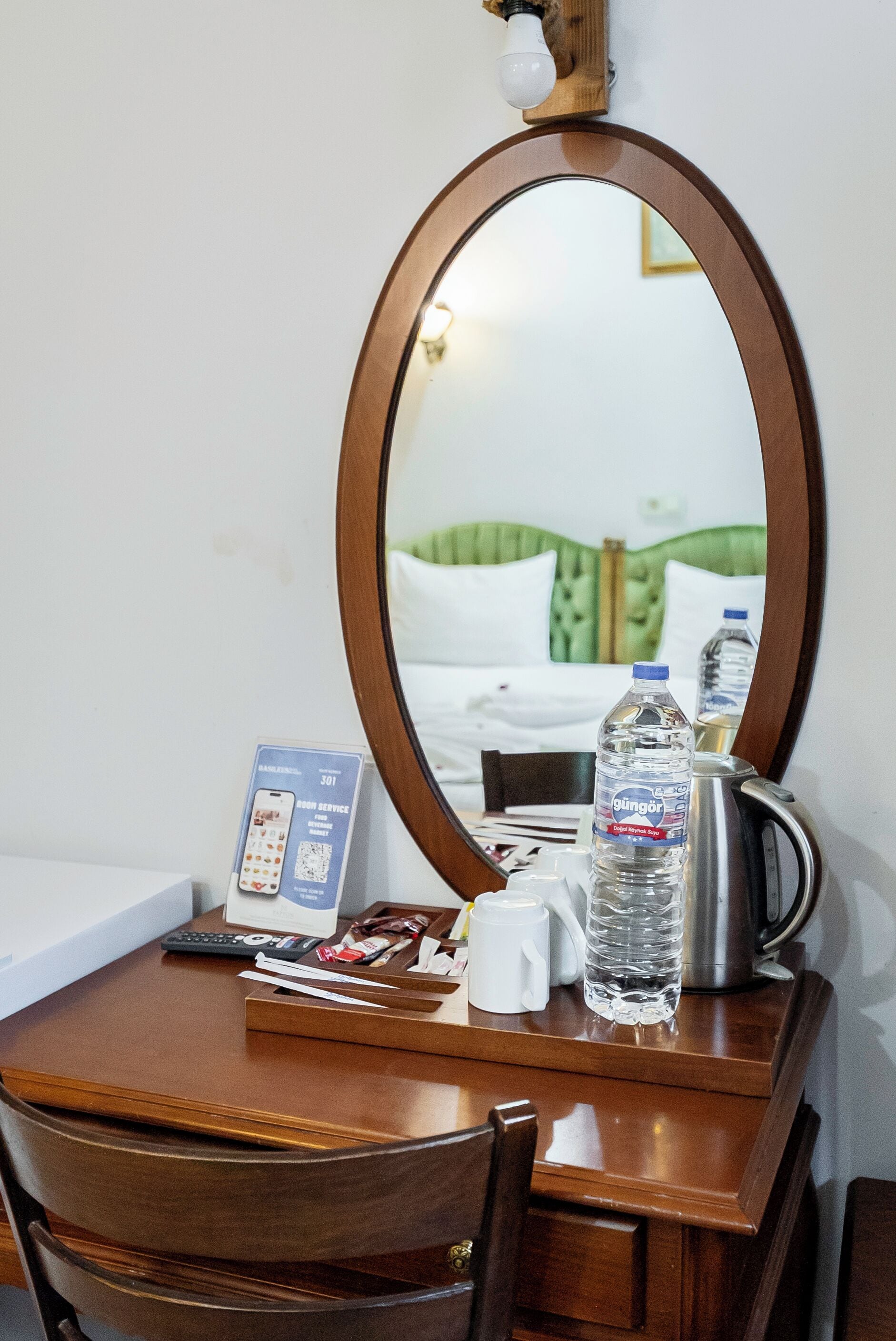 A hotel room with a wooden desk, a mirror, a telephone, a water bottle, a kettle, and a bed with green bedding.