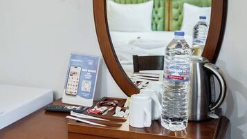 A hotel room with a wooden desk, a mirror, a telephone, a water bottle, a kettle, and a bed with green bedding.