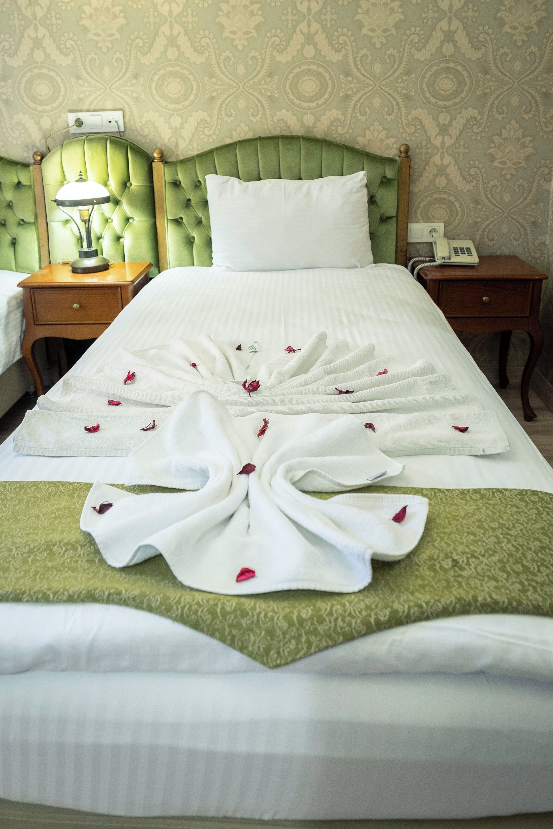 A neatly made bed with a green bedspread and a white duvet, a green headboard, a bedside lamp, and a telephone on the nightstand.