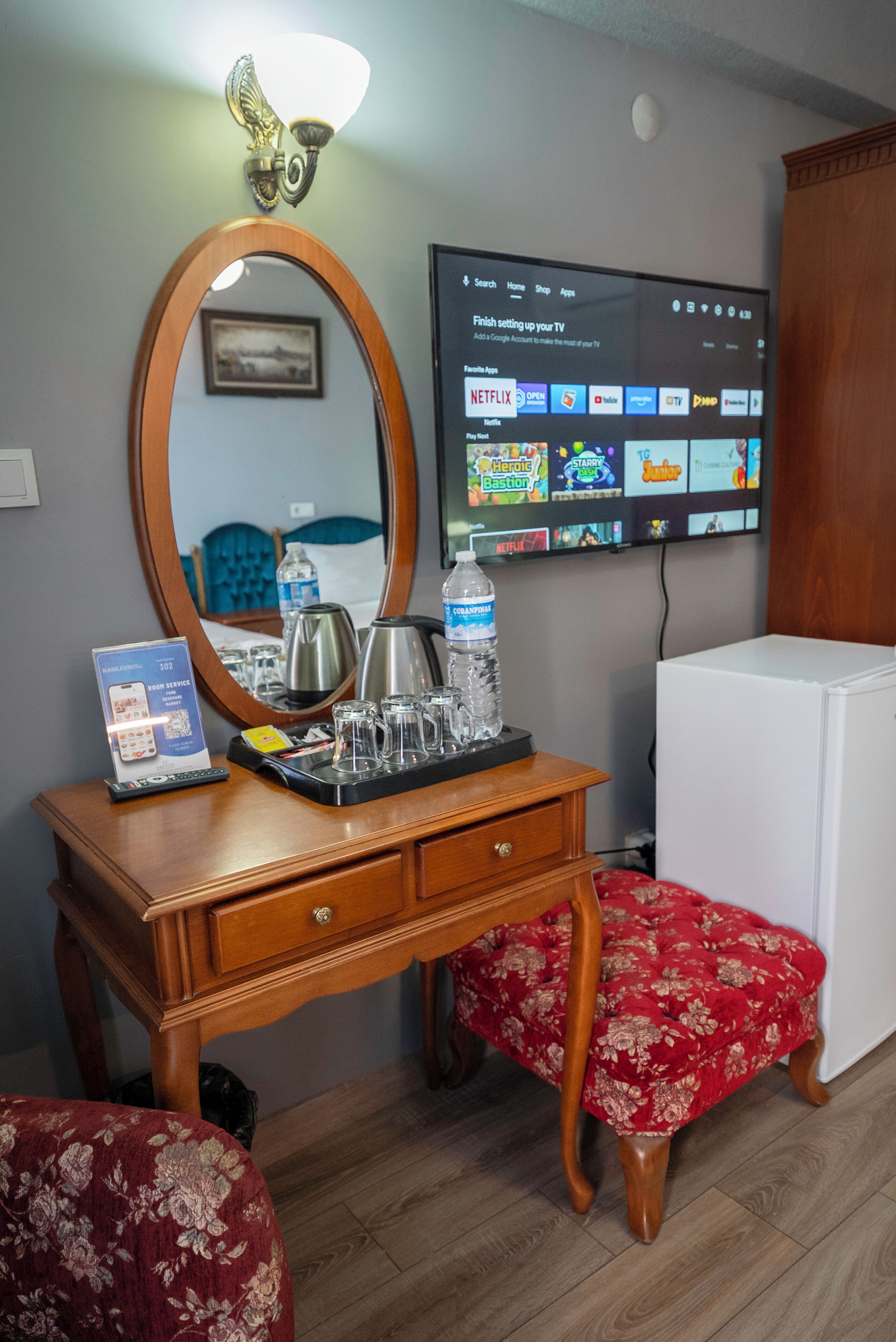 A hotel room with a wooden dresser, a mirror, a flat-screen TV displaying a TV menu, a water bottle, and a remote control.