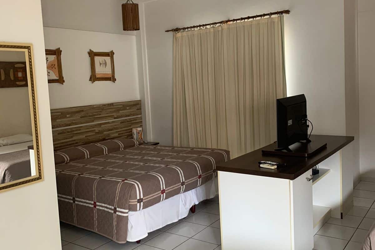 A hotel room with a bed, a desk with a computer, a mirror, and a window with curtains.