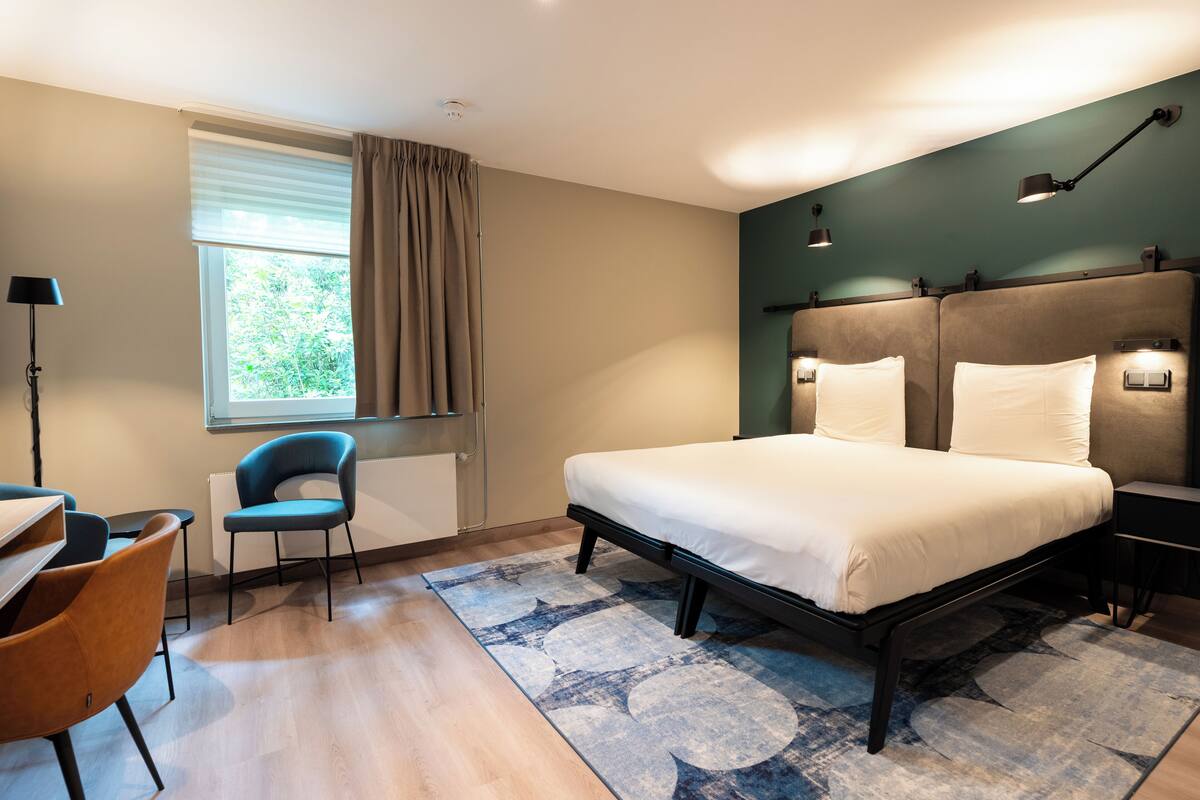 A modern hotel room with a large bed, a desk with a chair, a window with curtains, and a nightstand with a lamp.
