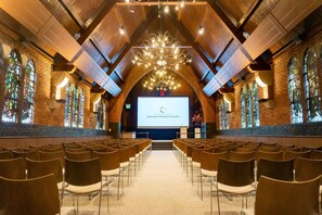 A conference room with a large screen at the front, rows of chairs, and stained glass windows. - Conferentiehotel Kontakt der Kontinenten (Soesterberg)
