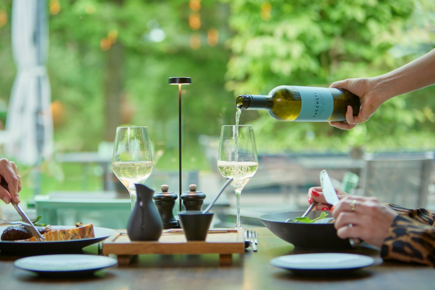 Una mesa de comedor con dos copas de vino, una botella de vino siendo servida, y una persona sosteniendo un tenedor.