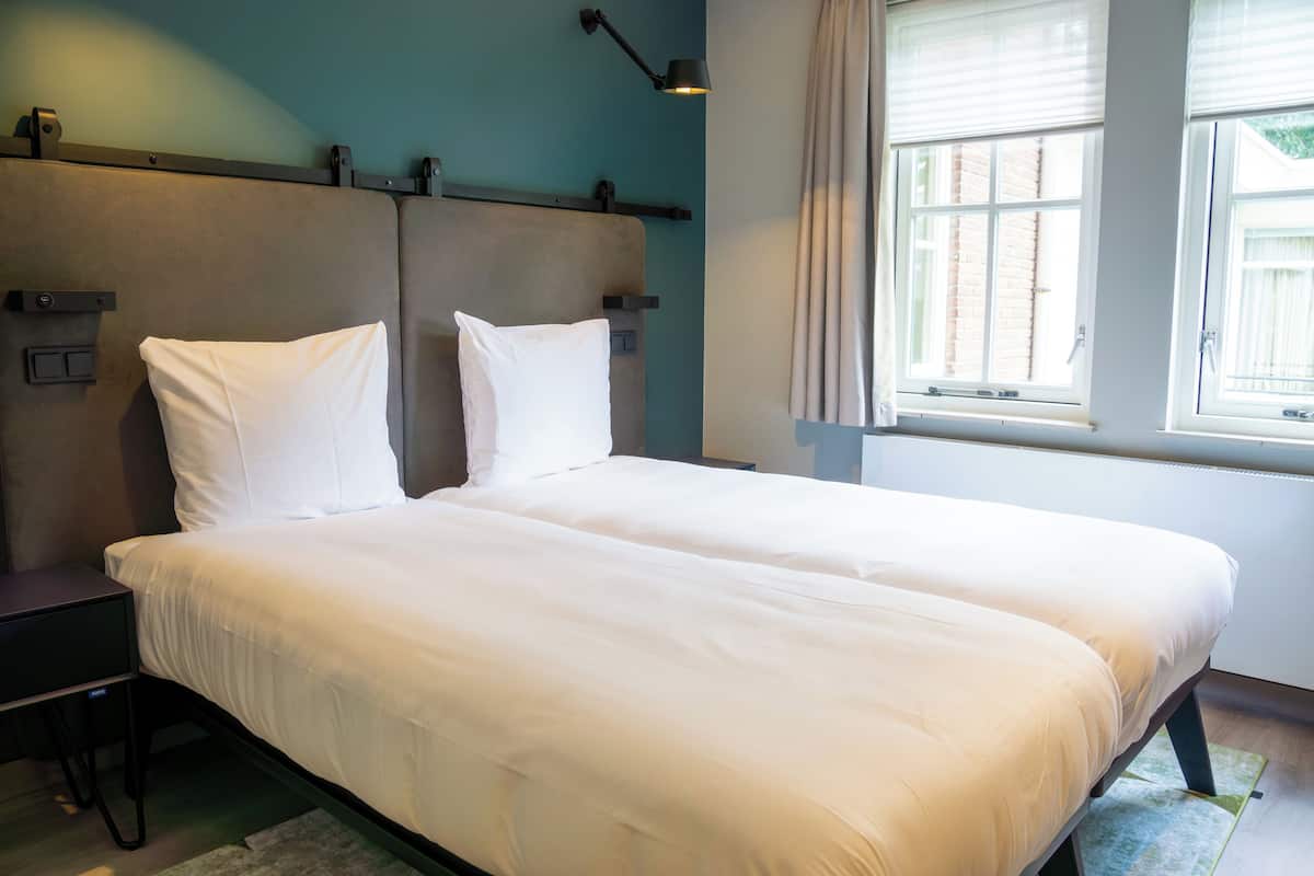 A double bed with white linens in a room with teal walls, a window with blinds, and a bedside table.