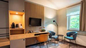 A modern hotel room with a wooden desk, a flat-screen TV, two chairs, a small table, and a window with a view of greenery. - Conferentiehotel Kontakt der Kontinenten (Soesterberg)