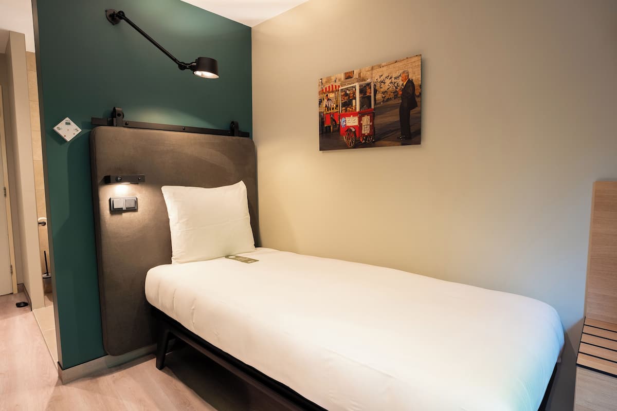A single bed with a headboard, a bedside lamp, a power outlet, and a wall-mounted picture.