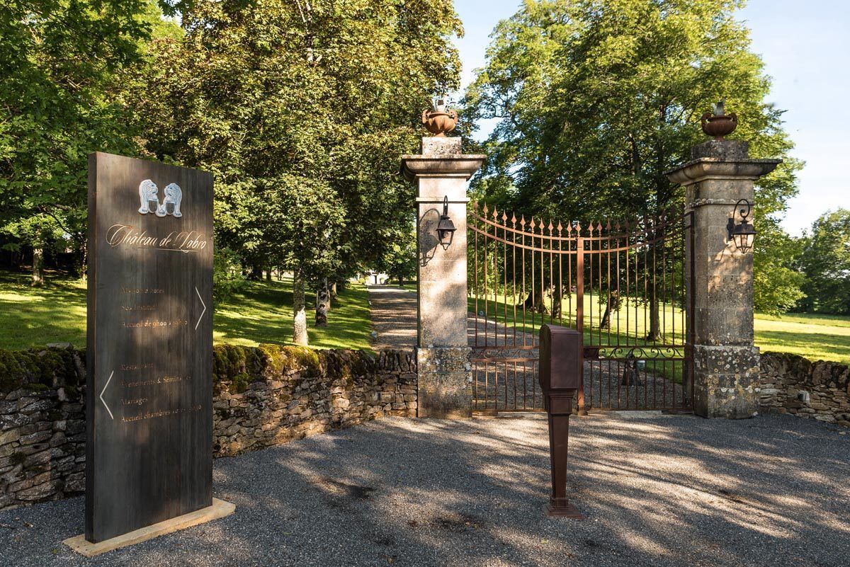 Une entrée fermée par un portail, agrémentée d’un panneau indiquant « Château de Labro », avec un mur en pierre et un chemin de gravier.