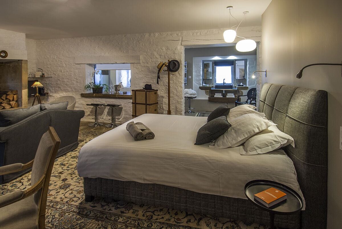 A hotel room with a large bed, a chair, a small table, a mirror, and a view of the outside.
