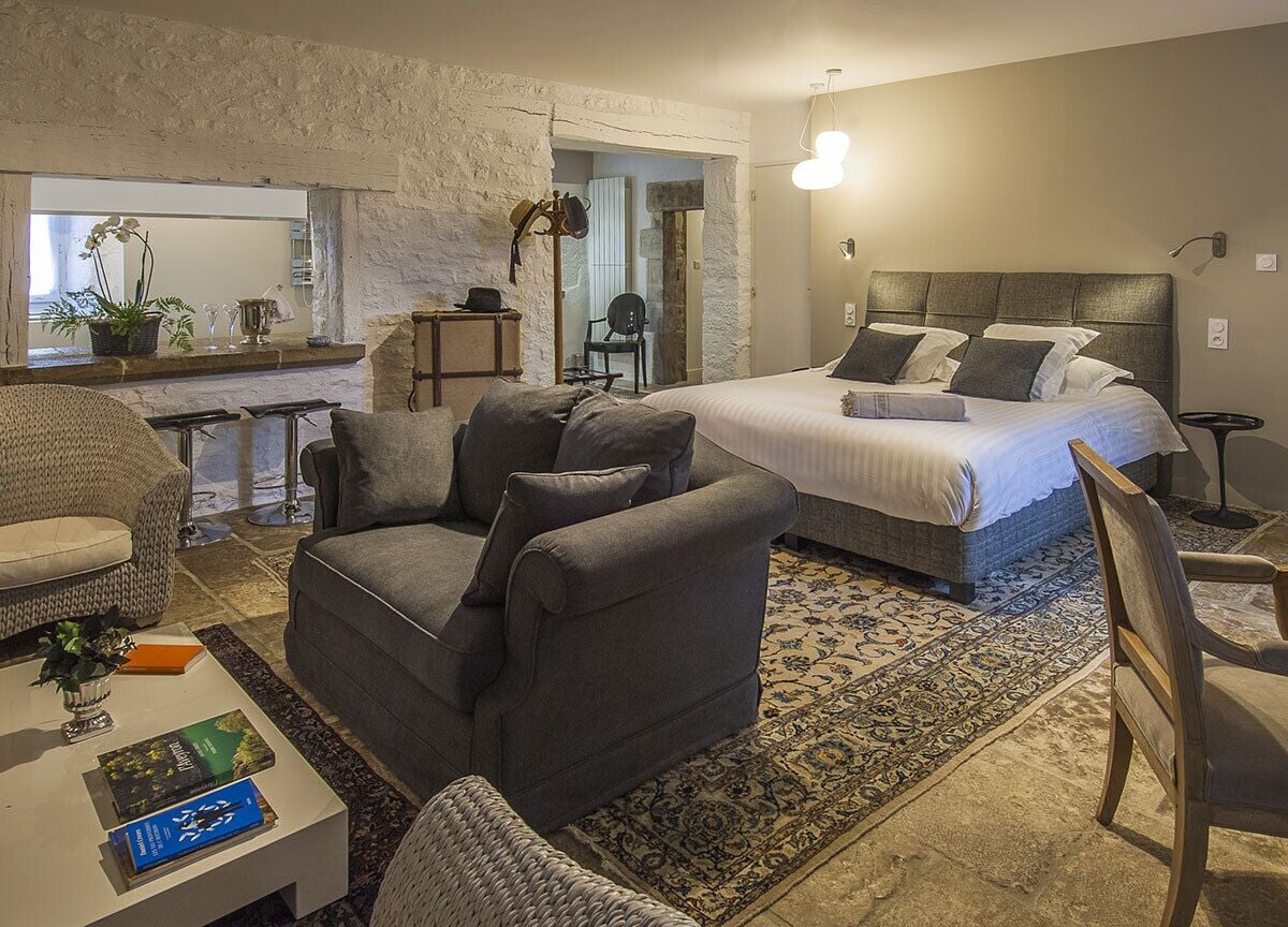 Une chambre moderne avec un grand lit, un coin salon comprenant un canapé et des fauteuils, une table basse avec des livres, et une vue sur la cuisine par une porte ouverte.