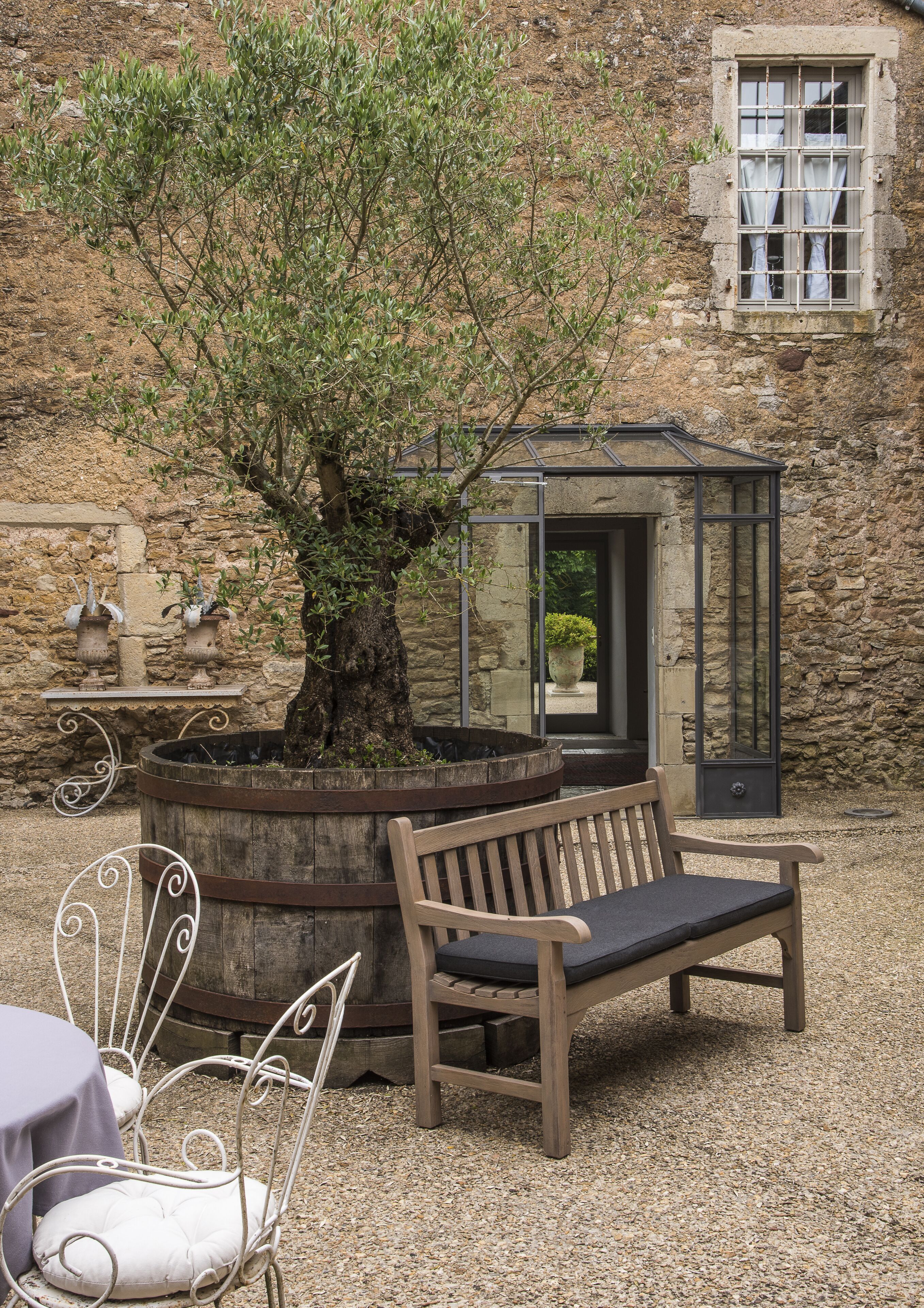 A rustic outdoor seating area with a wooden bench and metal chairs, a stone wall, and a small garden with a tree and potted plants.