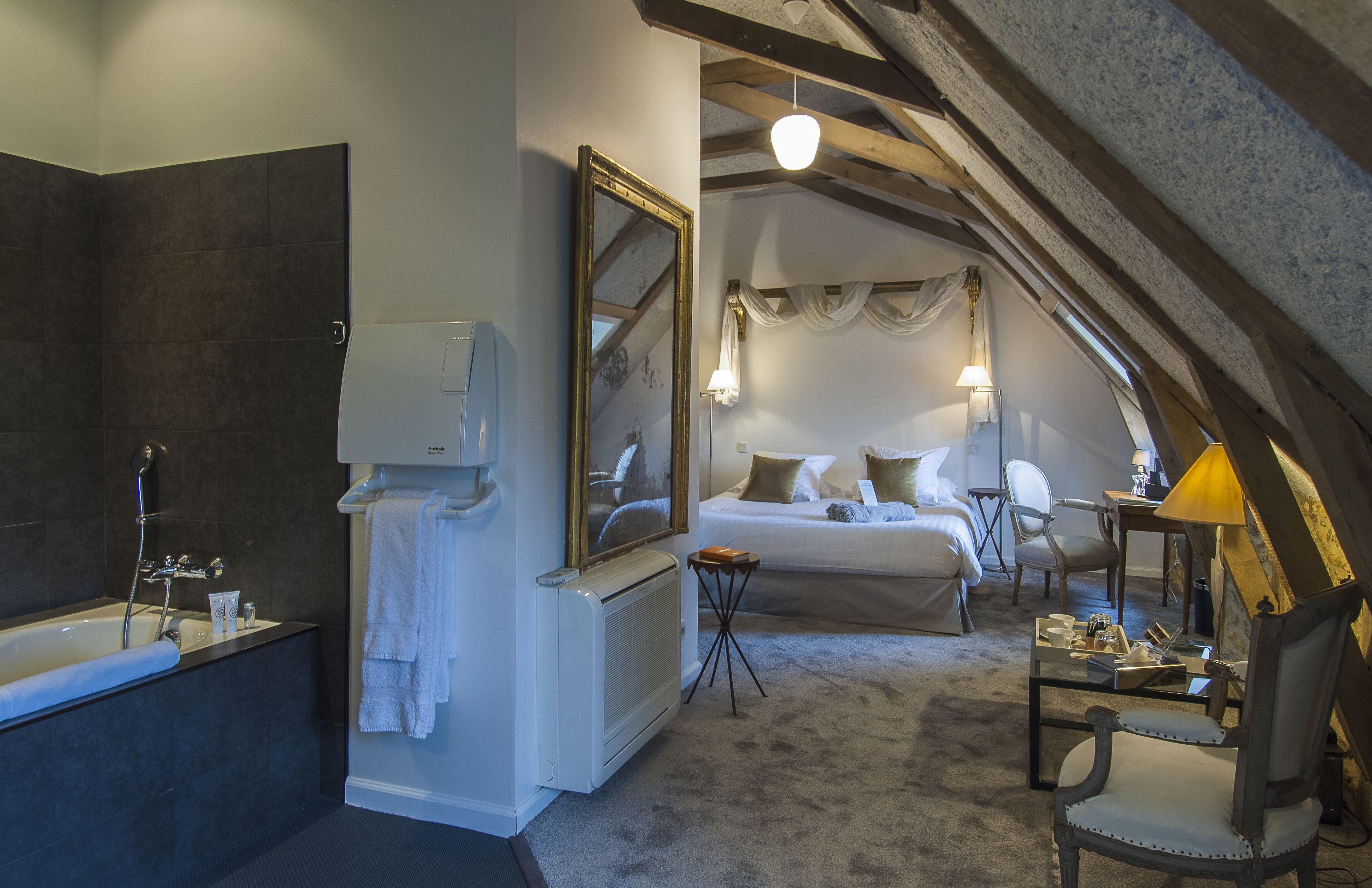 Une chambre dotée d’un grand lit, d’un coin salon avec un fauteuil et d’une salle de bain avec une baignoire et un miroir.
