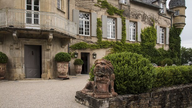 A historic building with a stone facade, multiple windows with shutters, a balcony with a wrought iron railing, and a statue in the foreground.