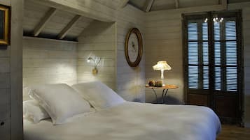 Une chambre avec un plafond en bois, un lit recouvert de draps blancs, une lampe de chevet, un miroir encadré et une fenêtre avec des stores.