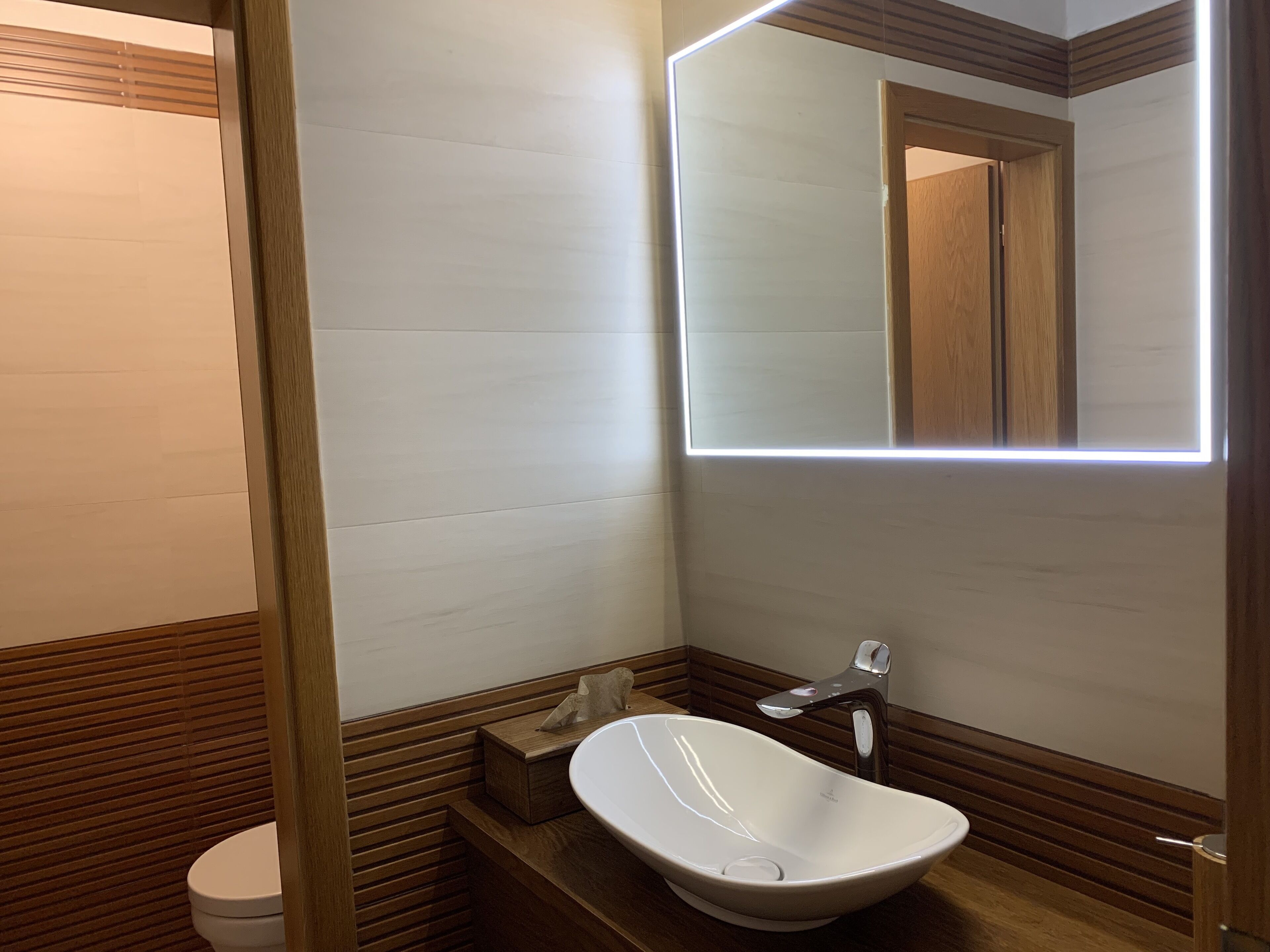 A modern bathroom with a wooden vanity, a white sink, and a mirror with integrated lighting.