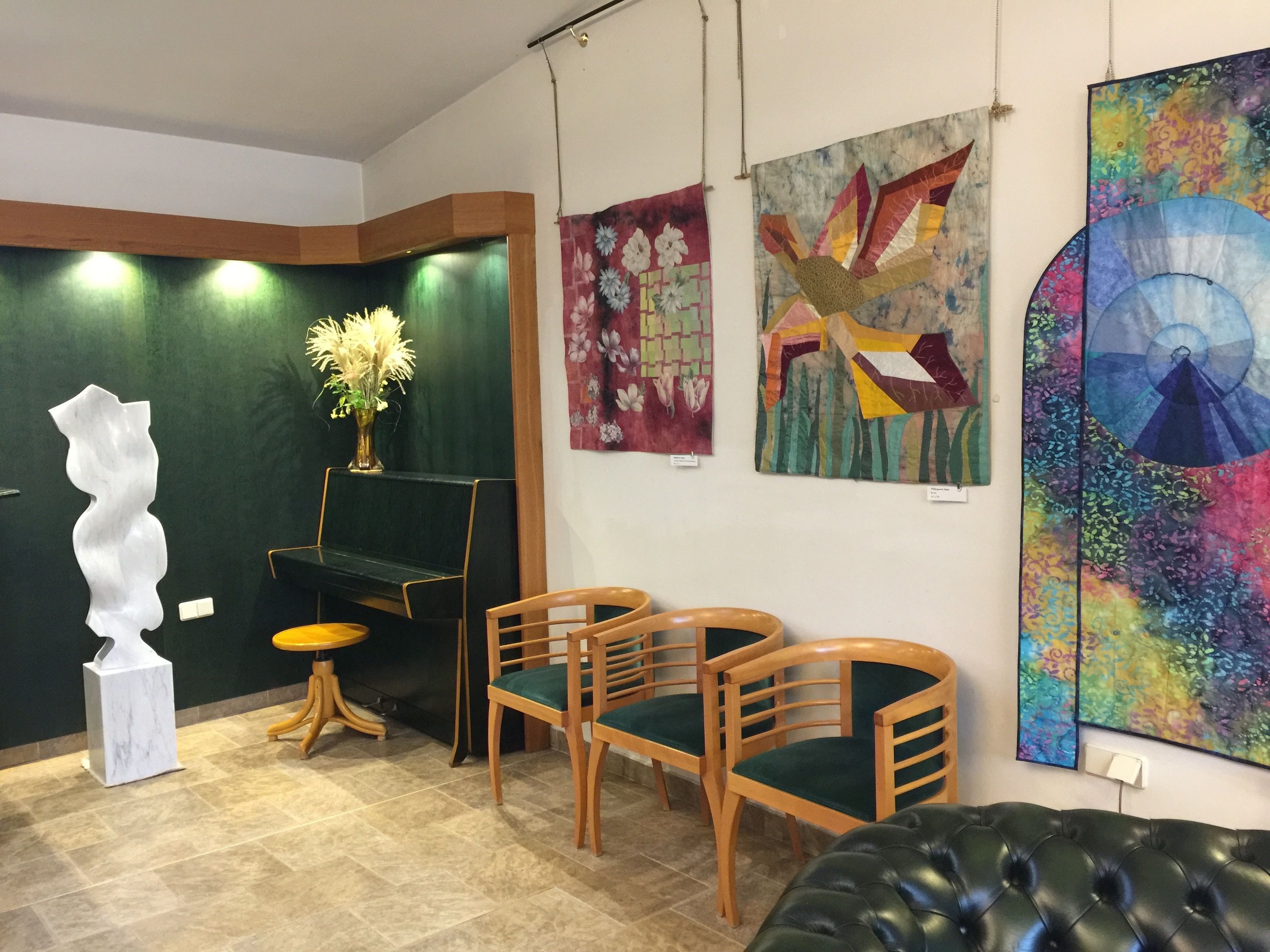 Een kamer met een groene wand, een witte abstracte sculptuur, een houten bank en drie houten stoelen met groene kussens.