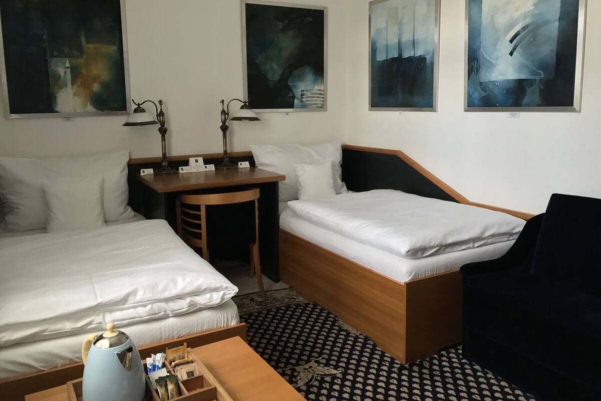 Ein Hotelzimmer mit zwei Einzelbetten, einem Schreibtisch mit zwei Lampen, einer Kaffeemaschine und drei abstrakten Bildern an der Wand.