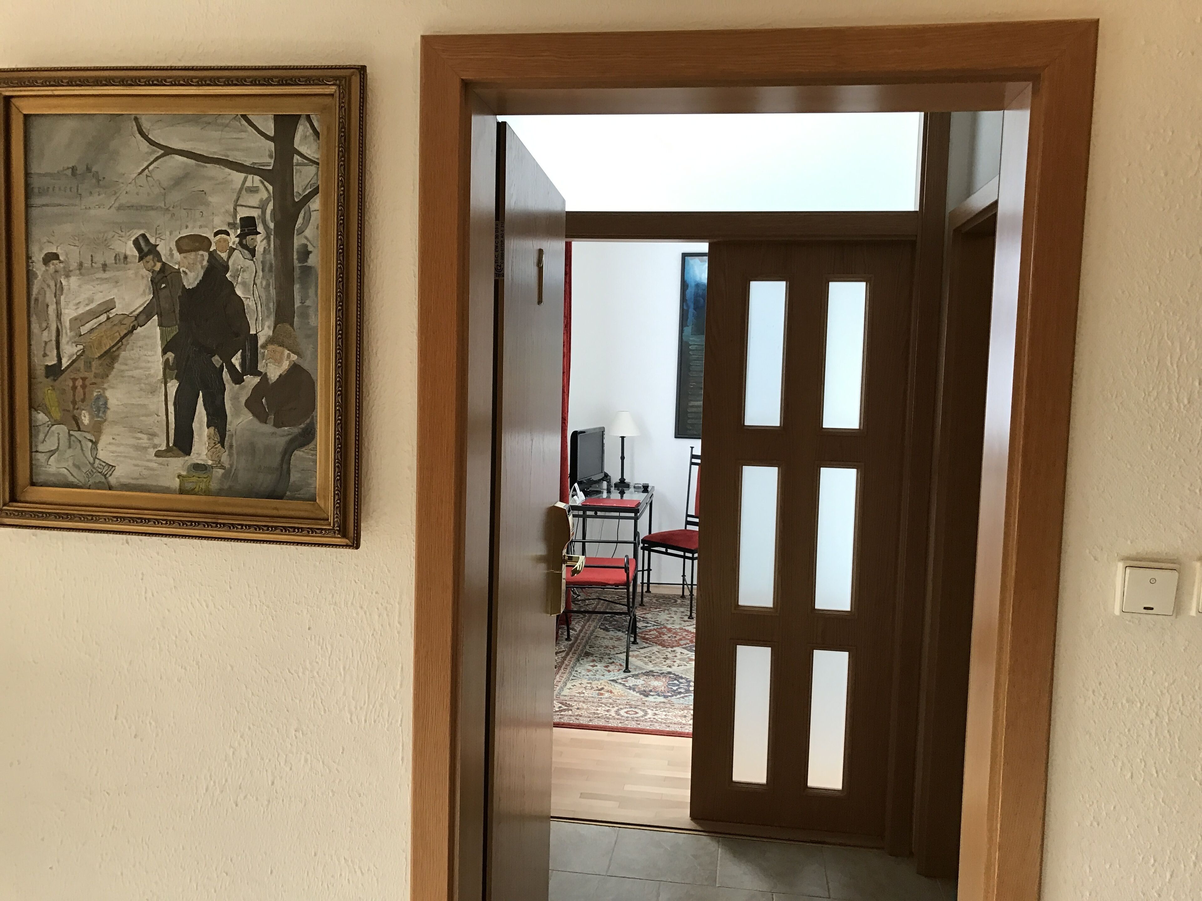 A hallway with a wooden door, a framed painting, and a glimpse of a room with a desk and chair.