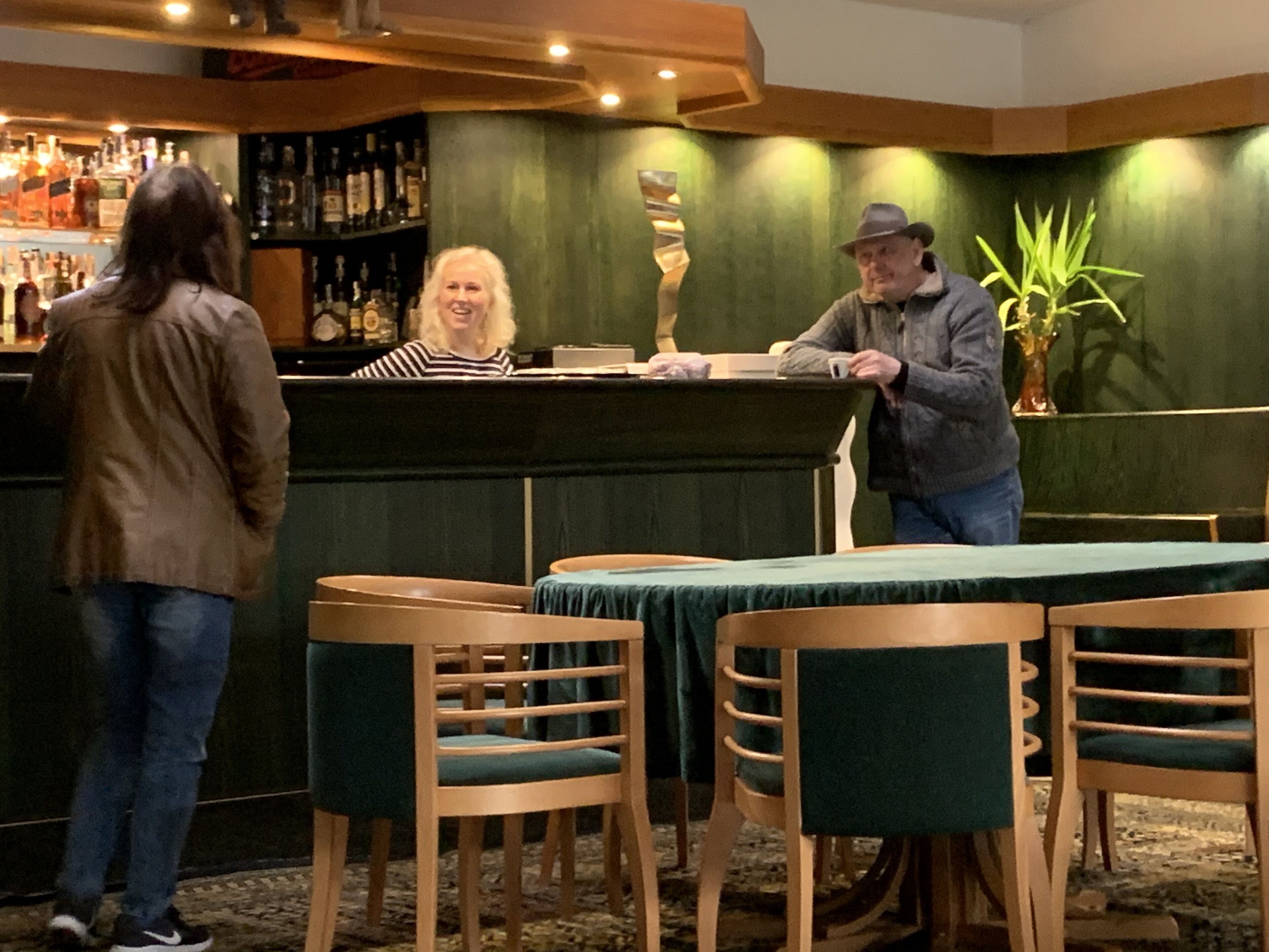 A bar area with wooden chairs and a green tablecloth, a bar counter, and a shelf with bottles.