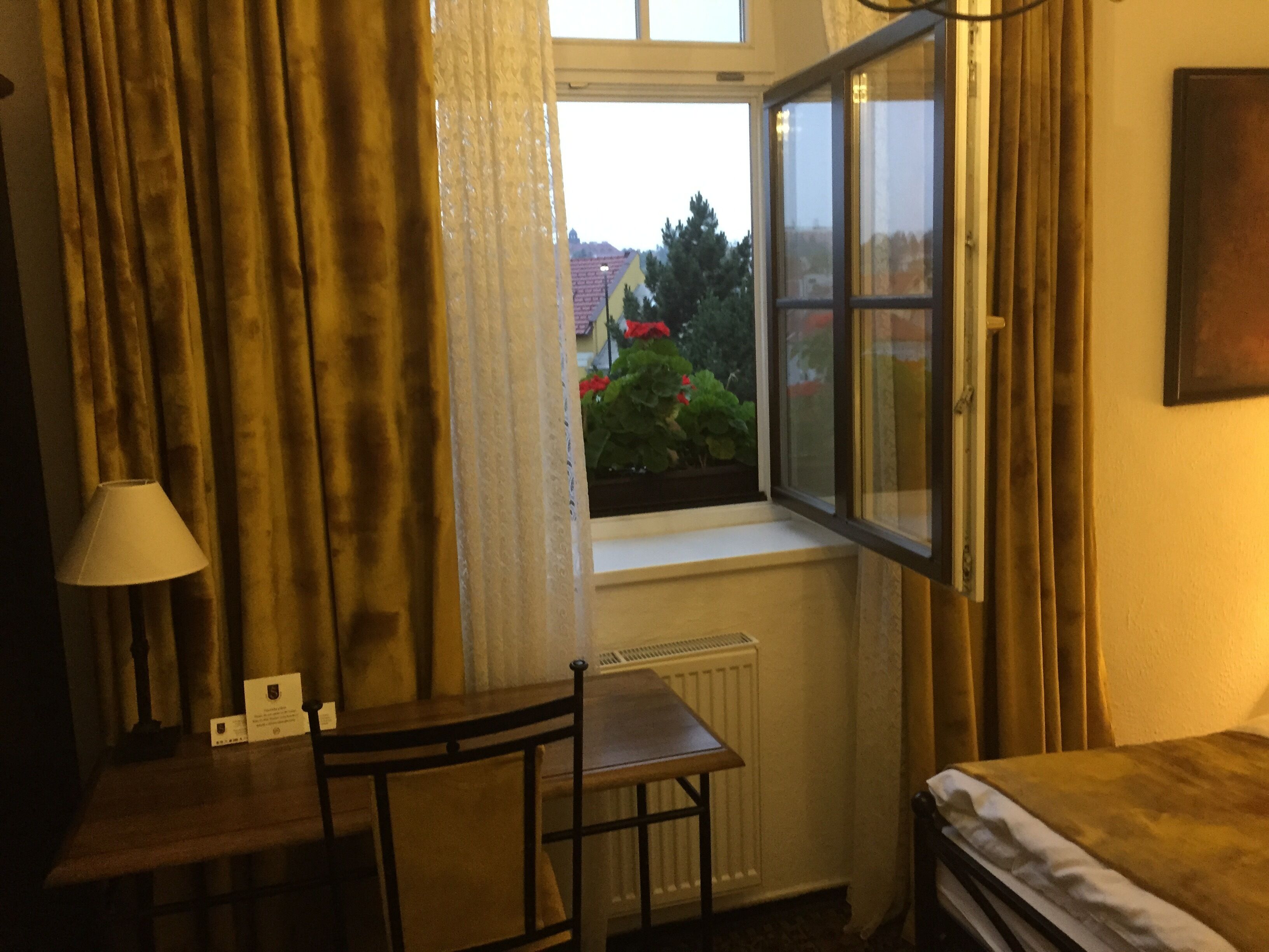 A hotel room with a bed, a small table, a chair, a window with curtains, and a radiator.