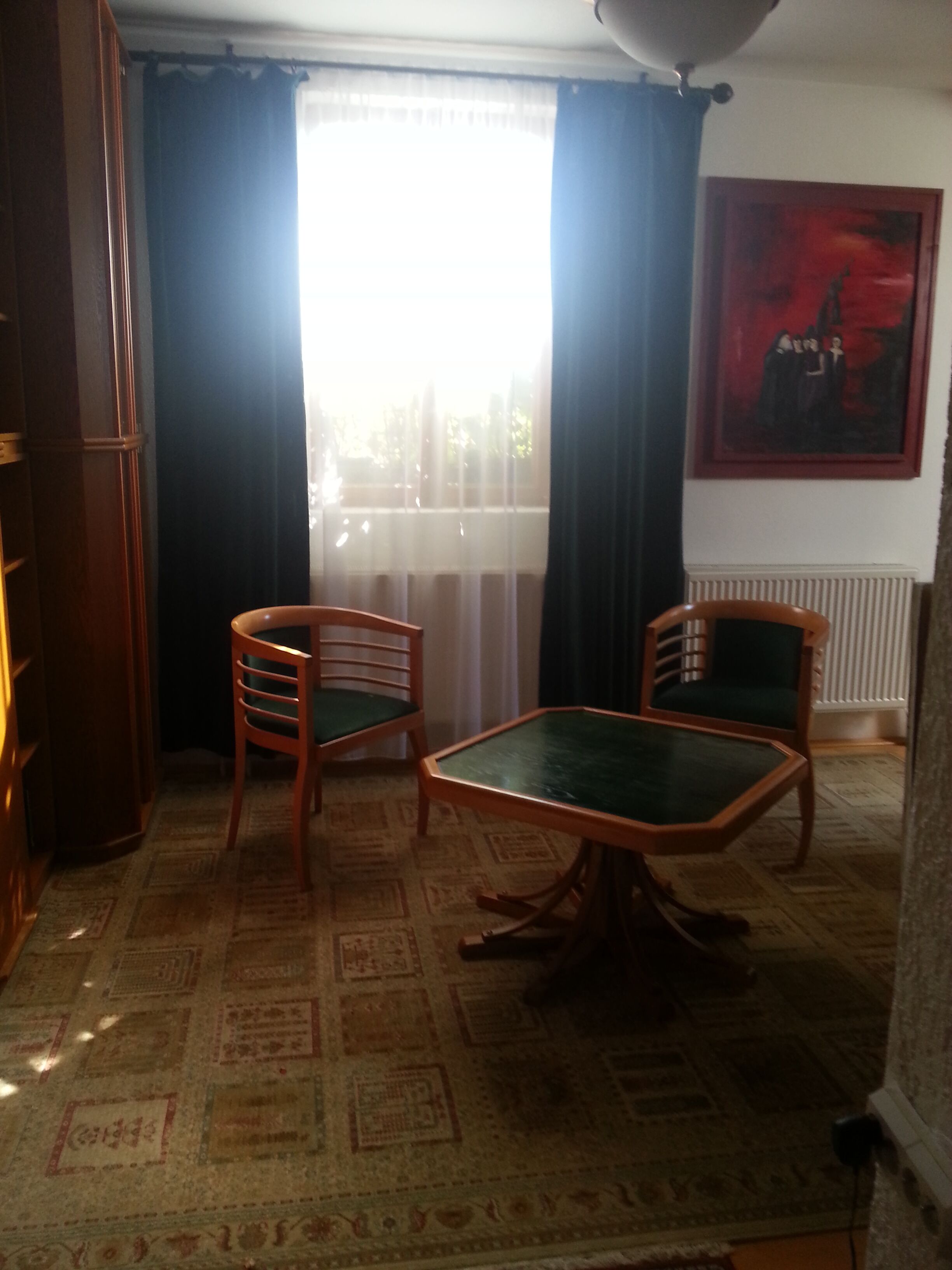 Een kamer met een kamerbreed tapijt, twee houten stoelen, een groene tafel, een radiator en een schilderij aan de muur.