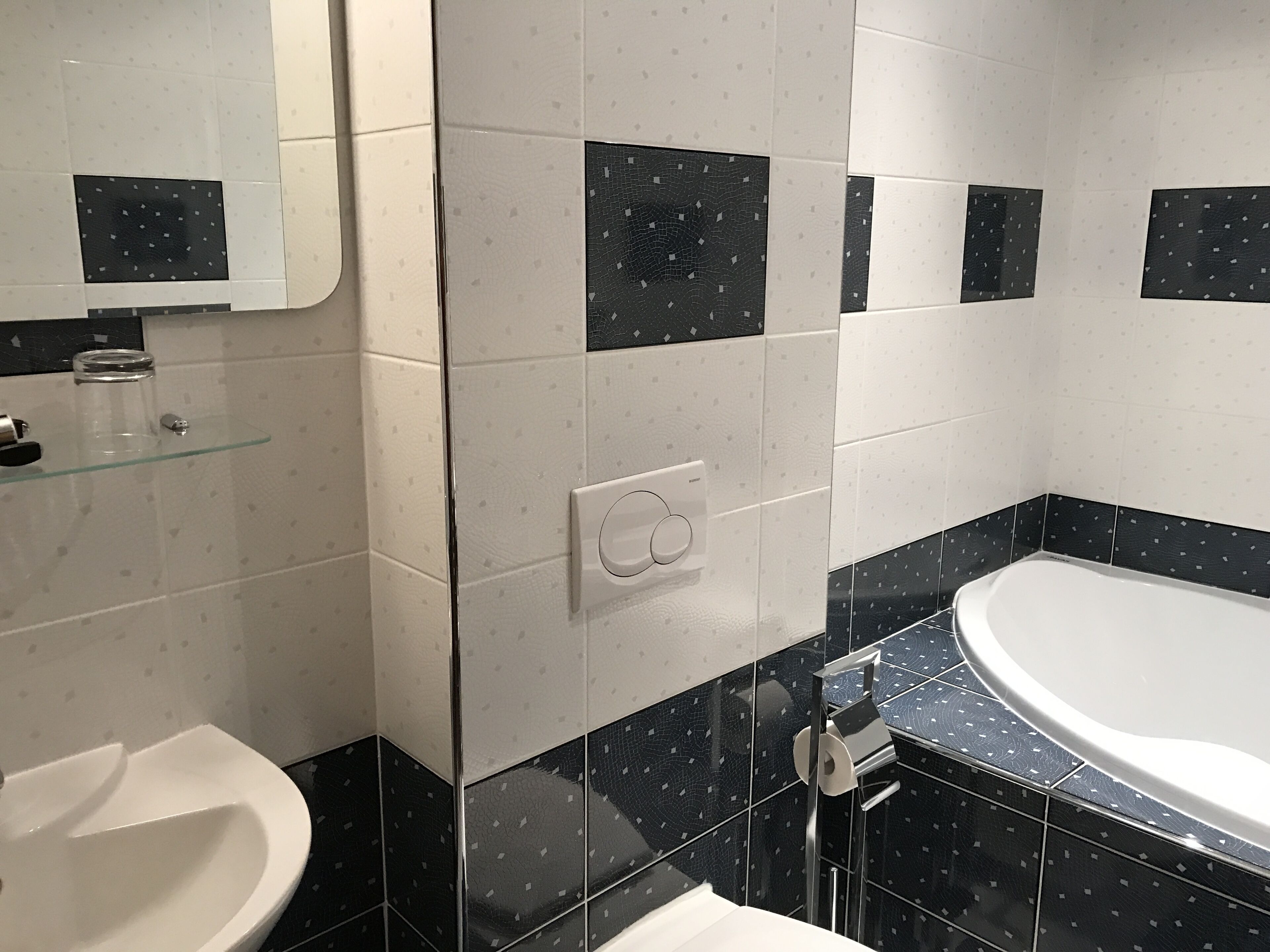 Un baño con un lavamanos blanco, un estante de vidrio, un espejo, una pared con baldosas blanco y negro y una bañera blanca.
