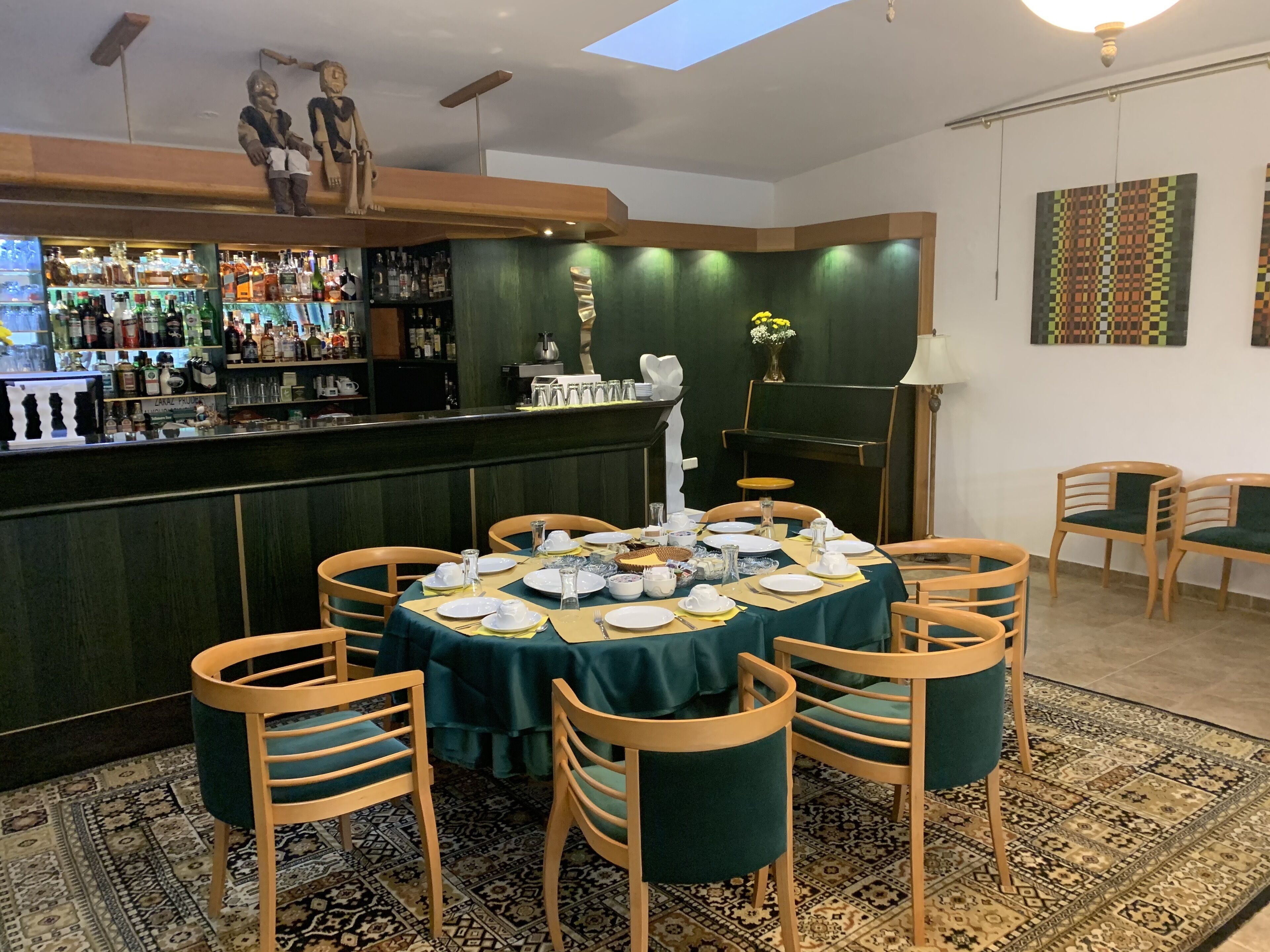 Un restaurante con zona de bar, sillas de madera, una mesa cubierta con mantel verde y una alfombra estampada.